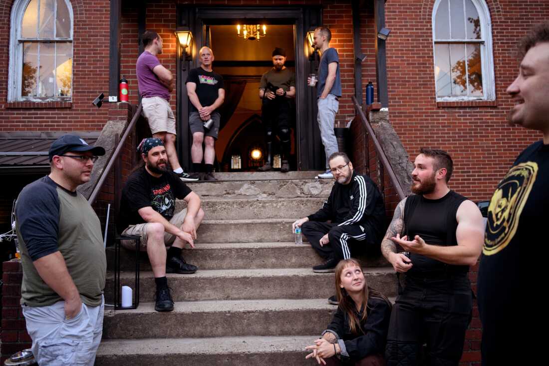 In this photo, nine men and one woman are congregated around the steps of the former church where the sword fights are held. They are wearing casual clothes. Some are sitting or standing on the steps, while a few are standing in front of the steps.