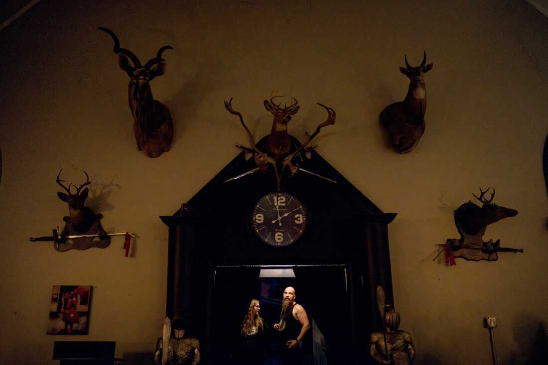 Chuck Gross stands in the doorway of the former Catholic church. He's wearing a dark tank top and has a long beard. Taxidermic animals with antlers are mounted on the wall above and around him. A teenage girl or young woman is to the left of him in the doorway.