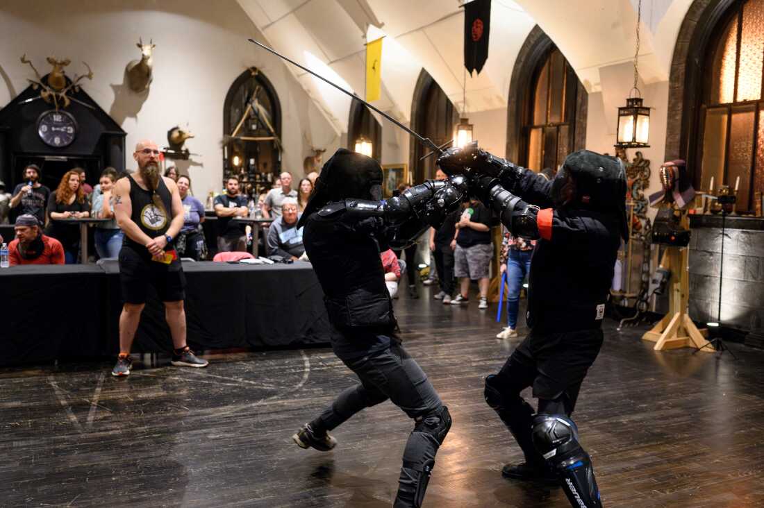 In this photo, two sword fighters, wearing all black and protective gear, fight against one another with long metal swords. In the background, audience members watch them compete in the tournament.