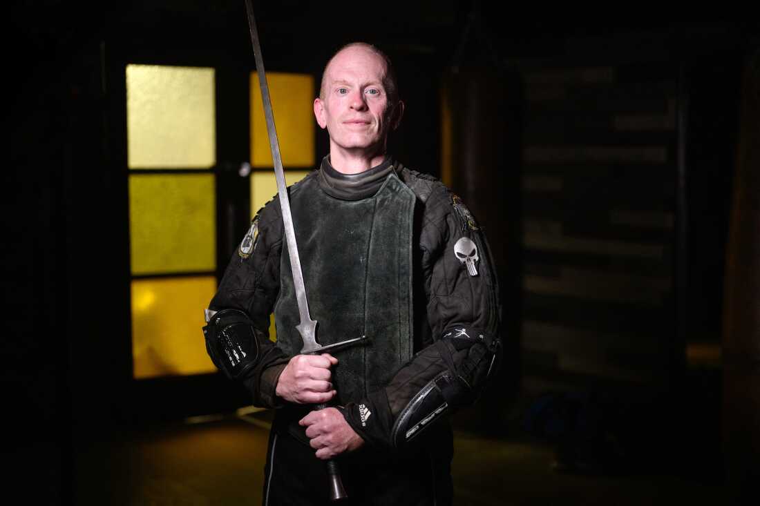 In this photo, Todd Rooney stands while holding a long metal sword. He's wearing a black protective sword-fighting outfit that has a skull patch on one sleeve.