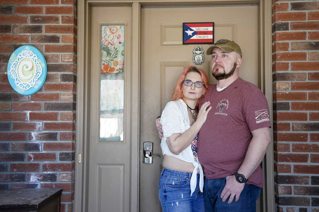 The Conlon hug as they stand outside the front door of their home. Their door has a sign that says "Welcome to the Colon's" painted on a Puerto Rican flag. They also have a blue sign with a decorative C hanging on the brick next to the door.