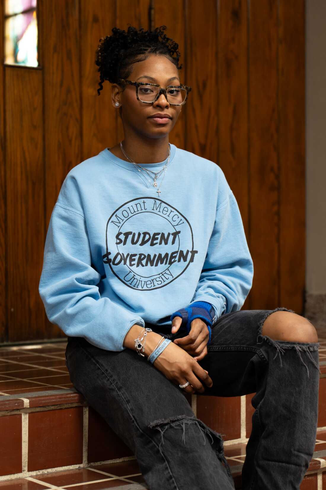 This photo shows Nasharia Patterson, Mount Mercy University's student government president. She's seated indoors on steps and is wearing dark jeans and a light blue sweatshirt. She has a brace on one wrist from an awkward back-tuck basket catch during cheer practice.