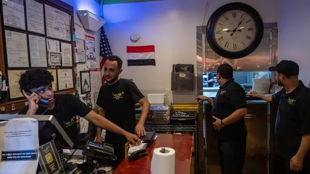  4 أشخاص يعملون في مقهى ومطعم "يمني" في الولايات المتحدة، يرتدون قمصاناً سوداء قصيرة الأكمام، أحدهم يتحدث على الهاتف. 8 يونيو/حزيران 2025 في منطقة بروكلين في مدينة نيويورك.