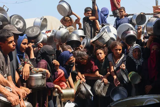 مجموعة من الأطفال والنساء يحملون أواني طعام فارغة ينتظرون على أمل الحصول على طعام. الصورة التقطت نهاراً.