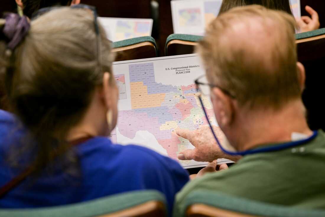 Photographed from behind, two members of the public, a man and a woman, look at a proposed redistricting map of Texas that they are holding in front of them. The man is pointing to a location on the map. The two are seated at a public hearing in a Texas Senate committee in August 2025.