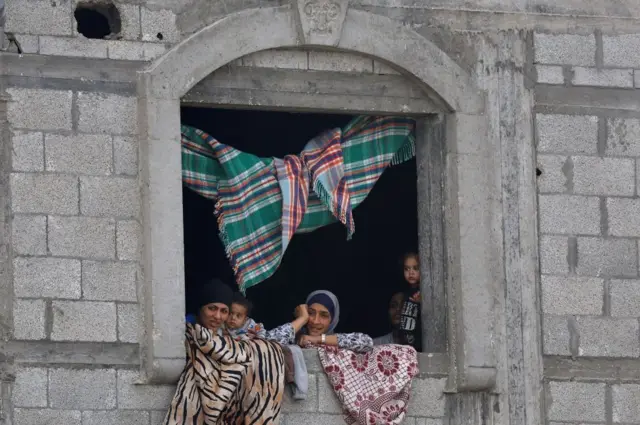 نساء وأطفال يطلّون من نافذة مبنى حجري. سجادٌ مُغطّى بالنافذة ومُعلّقٌ بالسقف.