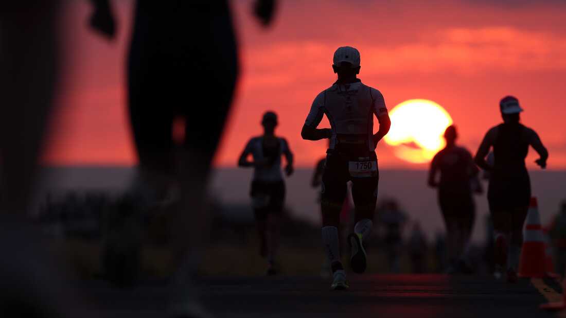 With a red sunset in the background, triathletes photographed in silhouette run in the final portion of the 2025 Ironman World Championship.