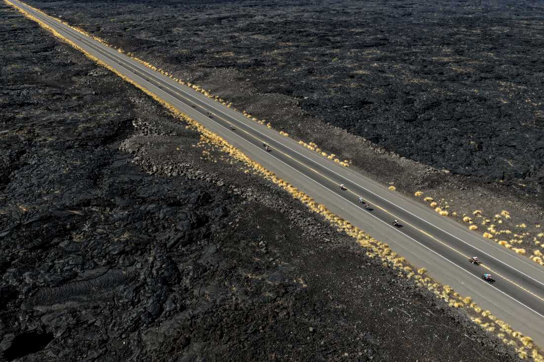 In this aerial photo, triathletes bike on a long straight road that cuts through a rough, black lava field during the Ironman World Championship on October 11, 2025, in Kailua-Kona, Hawaii.