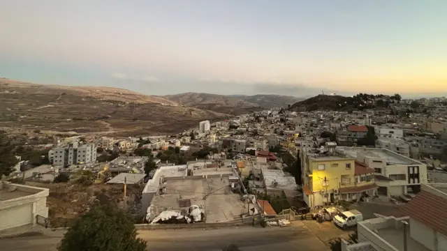 صورة من أعلى لبلدة صغيرة يظهر فيها الكثير من المباني الخرسانية المنخفضة. هناك تلال في الخلفية. السماء ملبدة بالغيوم لونها رمادي-أزرق، ويلوح في الأفق أثر خافت لأشعة الشمس.